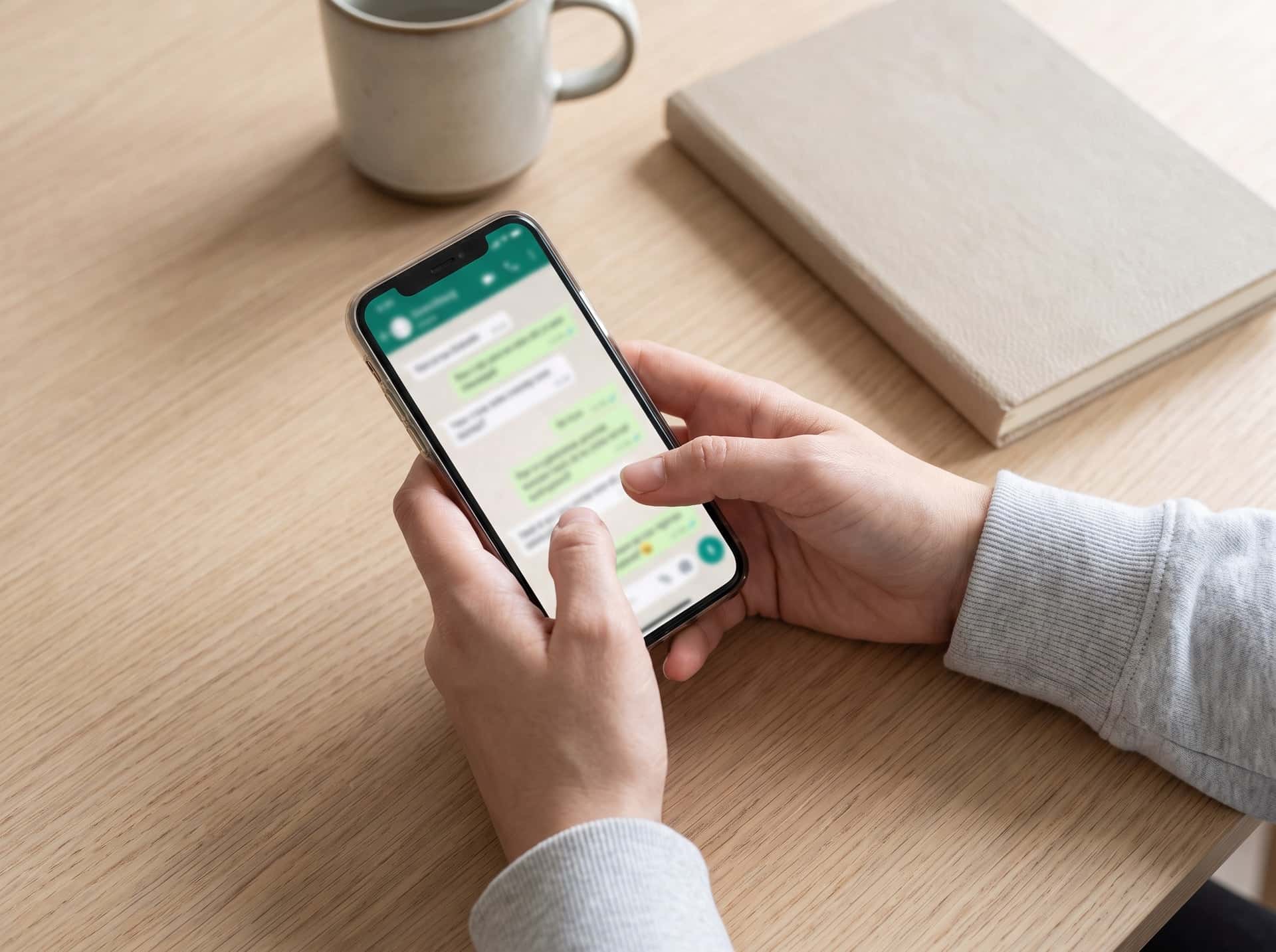 Mãos digitando uma mensagem no celular, sobre uma mesa
